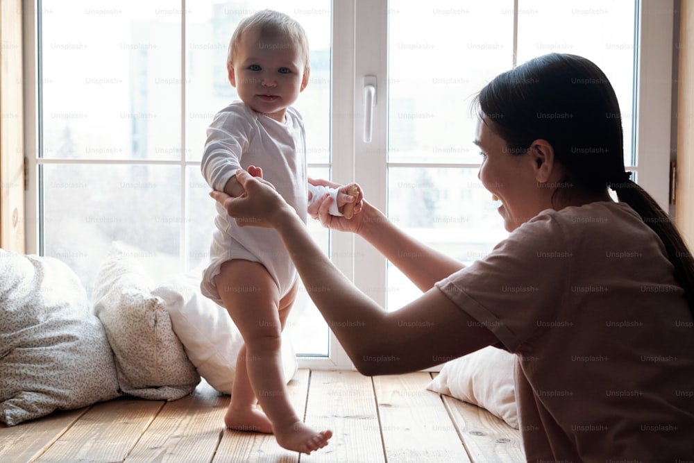 Fröhliche junge Frau, die ihr kleines Baby an den Händen hält und ihm hilft, erste Schritte zu machen