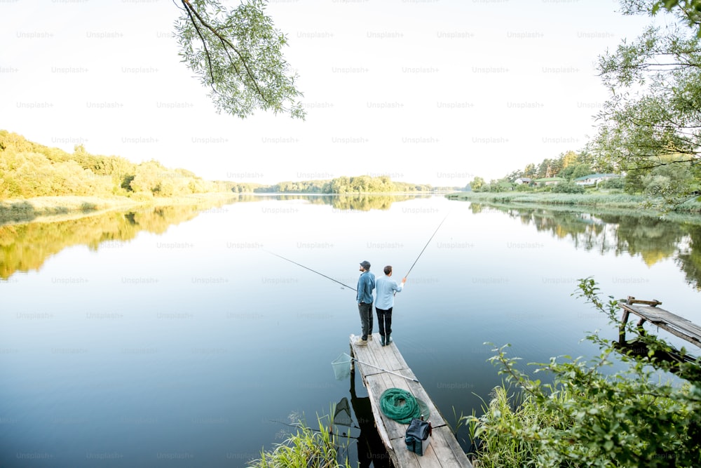 Landscape view on the beautiful lake with two male friends fishing together standing on the wooden pier during the morning light