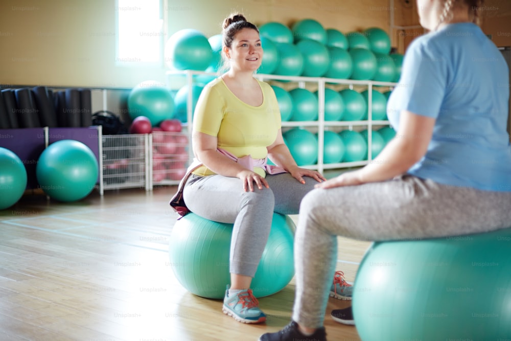 Two oversized young women sitting on fitballs in gym, talking and having workout