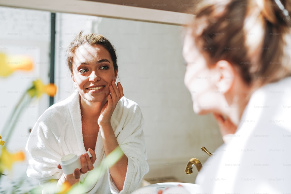 Portrait de beauté d’une jeune femme souriante aux cheveux longs foncés dans un peignoir blanc mettant une crème hydratante nourrissante de jour sur une peau propre et fraîche, le visage et les mains près du miroir dans la salle de bain à la maison