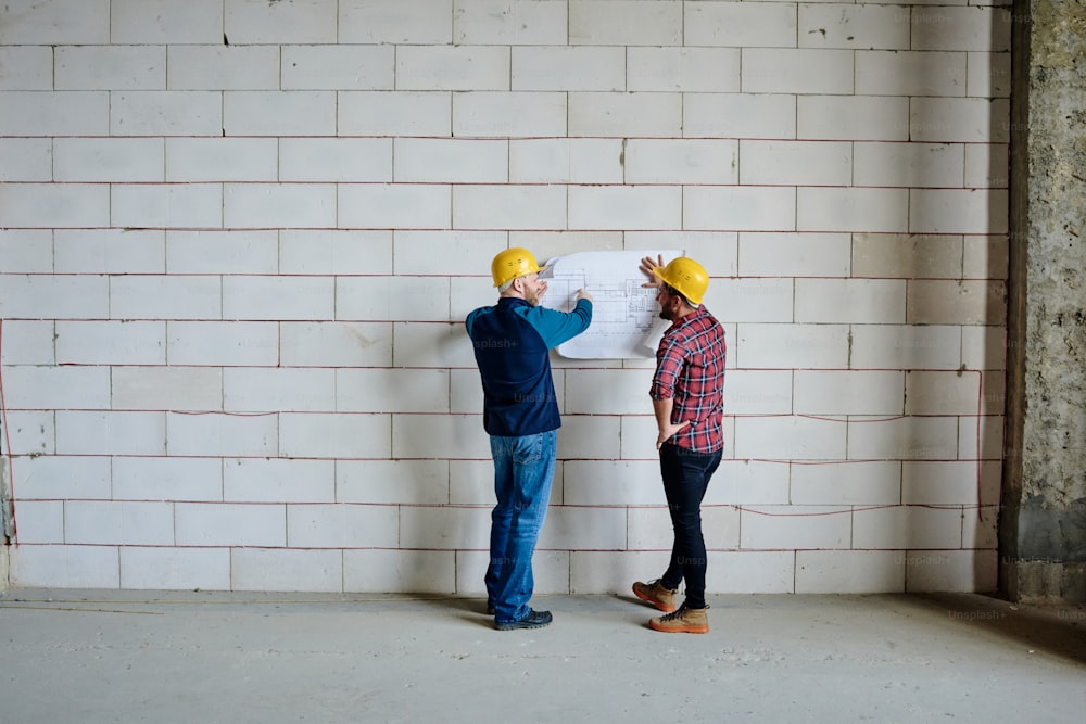 안전모를 쓴 두 명의 전문 엔지니어가 벽돌 벽 옆에 서서 작업 회의에서 미완성 건물의 스케치에 대해 논의하고 있습니다.
