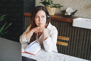 Donna bruna adulta cinquant'anni in cuffie che prende appunti nel libro quotidiano con laptop aperto, studia qualcosa con il corso online a casa