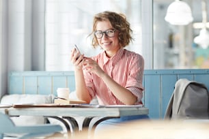 カフェのテーブルに座って、ソーシャルネットワークで友達とhehr写真を共有しながらカメラを見ている幸せな女の子