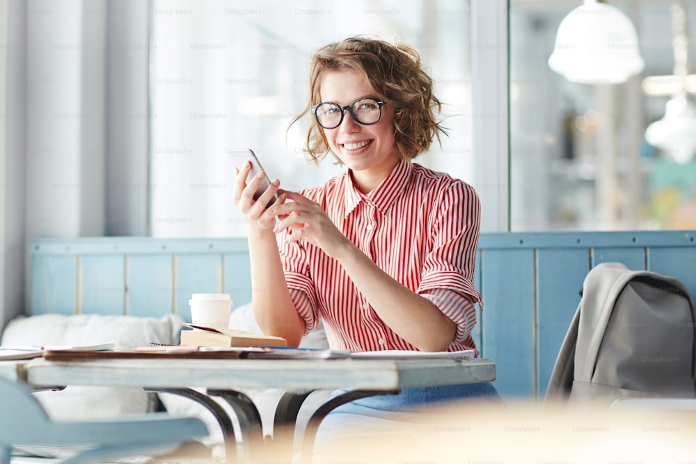 Glückliches Mädchen, das in die Kamera schaut, während es am Tisch im Café sitzt und Fotos mit Freunden in sozialen Netzwerken teilt