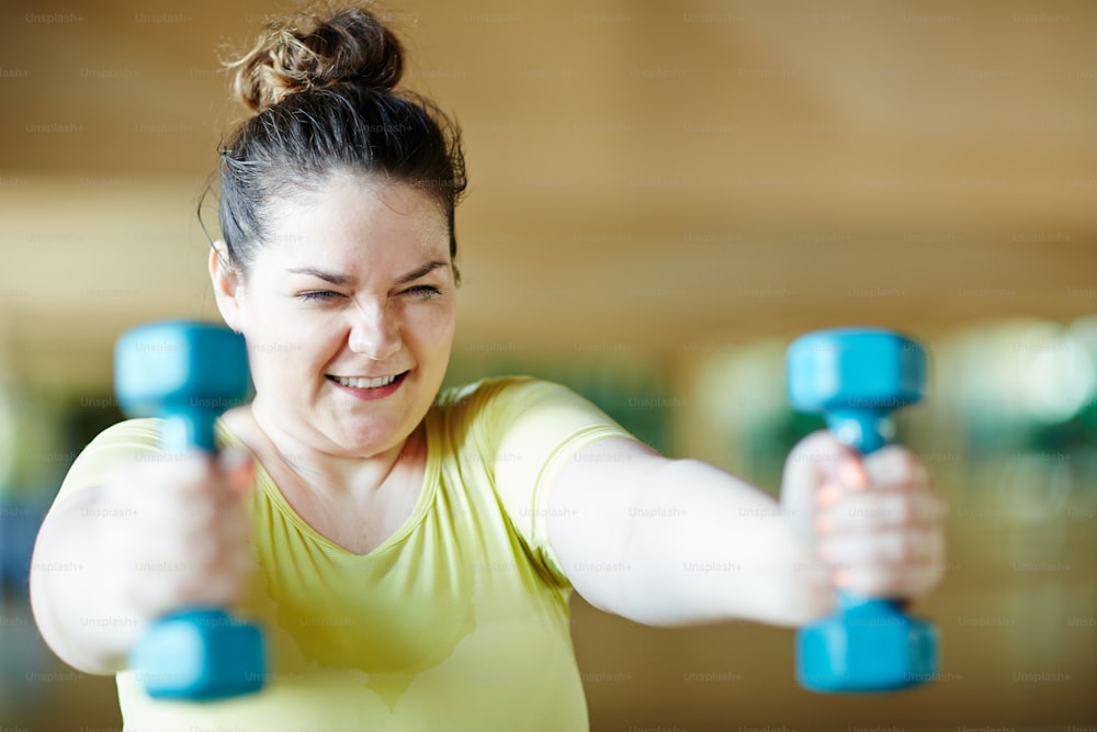 Chubby female in activewear sweating and working hard during training with dumbbells