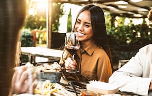 レストランのダイニングテーブルに座って赤ワインを飲む幸せなアジア人女性 - 外でバーベキューディナーパーティーをしている友人のグループ - 食べ物と飲み物のライフスタイルのコンセプト