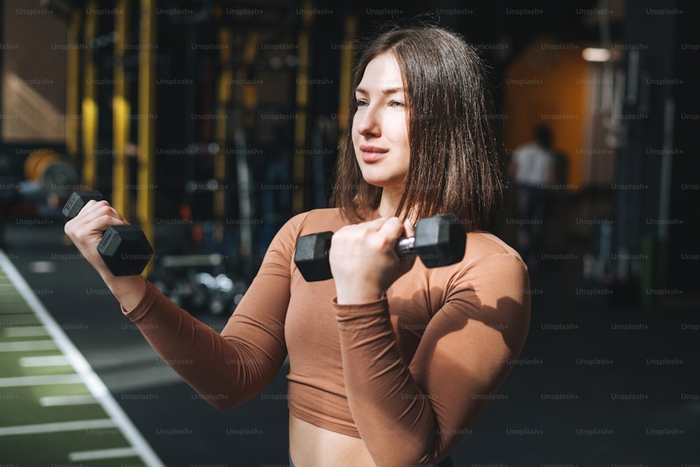Young brunette woman training her muscles with dumbbells in fitness club gym
