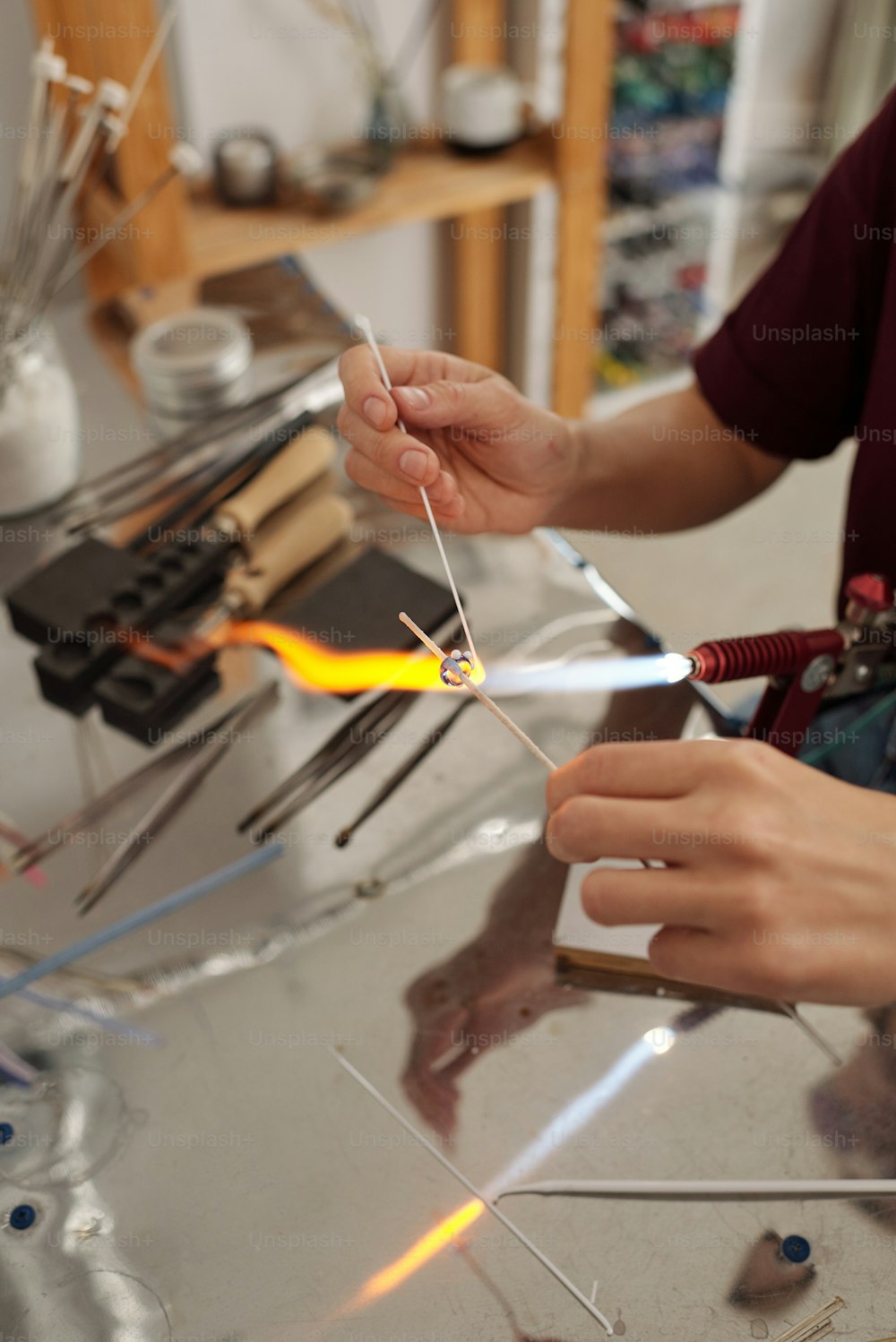 Hände einer jungen professionellen Lampenarbeiterin, die Glaswerkstück mit Feuer brennt, während sie es in Werkstatt oder Atelier über Brenner hält