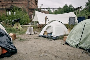 Campo de refugiados com tendas, roupas penduradas em corda e cadeiras dobráveis contra edifícios abandonados