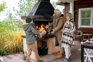 Happy senior couple in warm casualwear standing by fireplace of their country house and looking at grandson carrying box with firewood