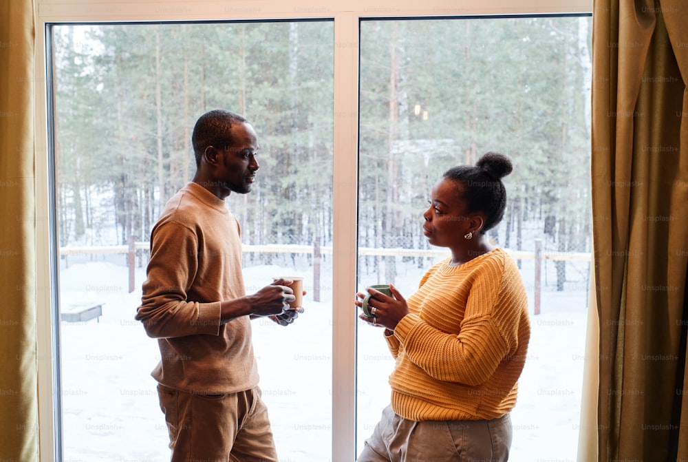Vista lateral de una joven pareja afroamericana en ropa informal tomando el té mientras están de pie uno frente al otro junto a la ventana de la casa de campo