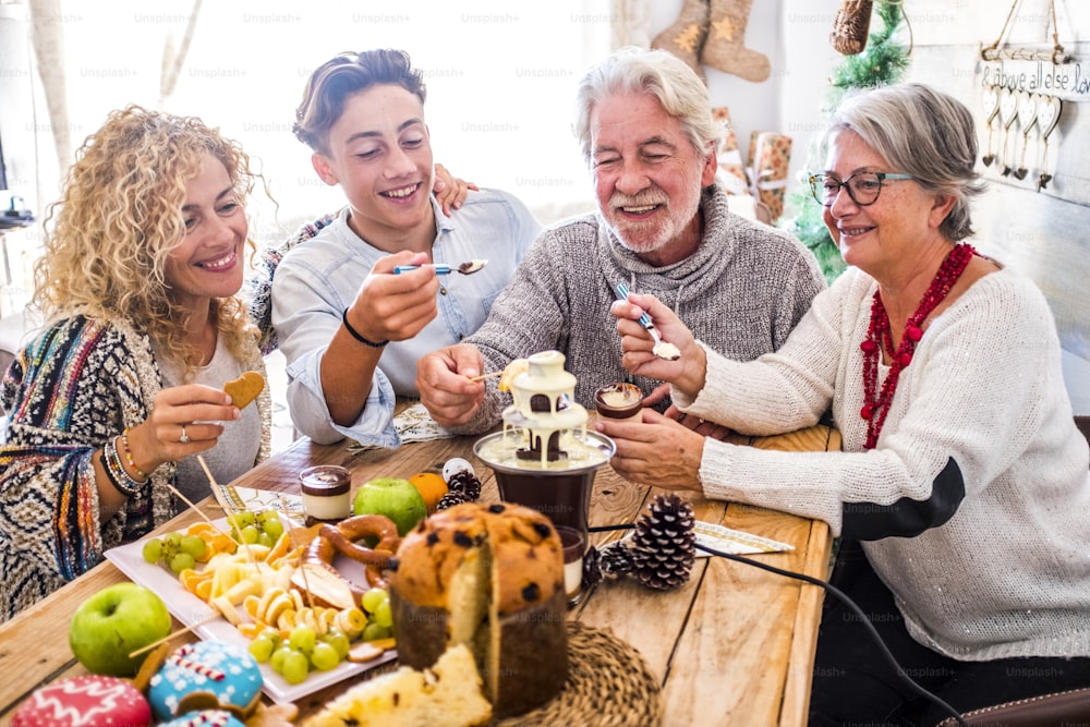 Cheerful family with mixed ages enjoy together the Christmas winter eve lunch at home with chocolate and cakes and frees - people celebrate event in friendship and love - white home indoor and wooden table