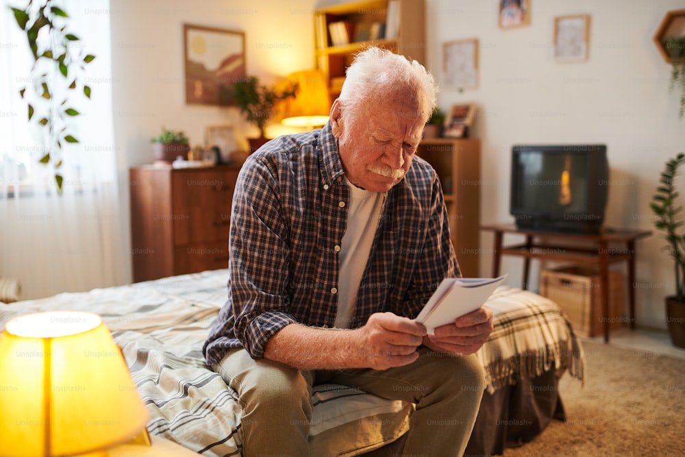 Contemporary senior man looking through photographs of his relatives while sitting on bed in home environment