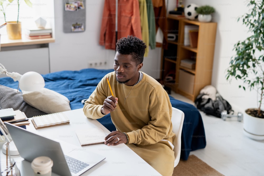 Joven estudiante africano en ropa de casa sentado frente a una computadora portátil y escuchando al maestro durante la lección en línea