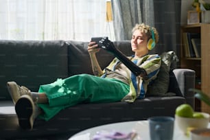Mädchen mit Behinderung in Kopfhörern, die Online-Filme auf dem Smartphone ansehen oder im Video-Chat kommunizieren, während sie sich auf der Couch im Wohnzimmer entspannen