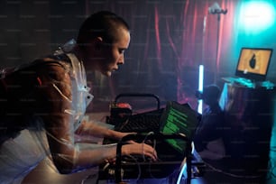 Jovem mulher séria em casaco de celofane decodificando dados e olhando para a tela do laptop contra o cyberpunk masculino sentado no chão