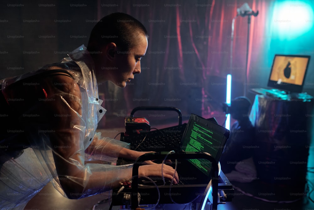Young serious female in cellophane coat decoding data and looking at laptop display against male cyberpunk sitting on the floor