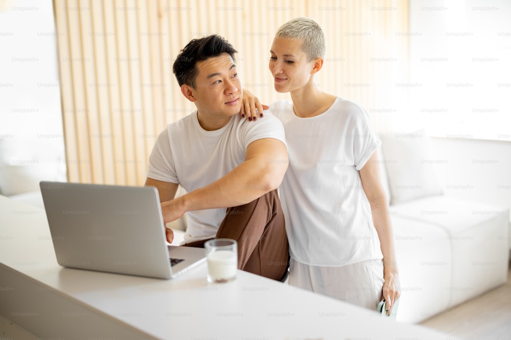 Caucasian woman watching something on laptop of her asian boyfriend. Concept of relationship and spending time together. Idea of domestic lifestyle. Multiracial couple in modern apartment