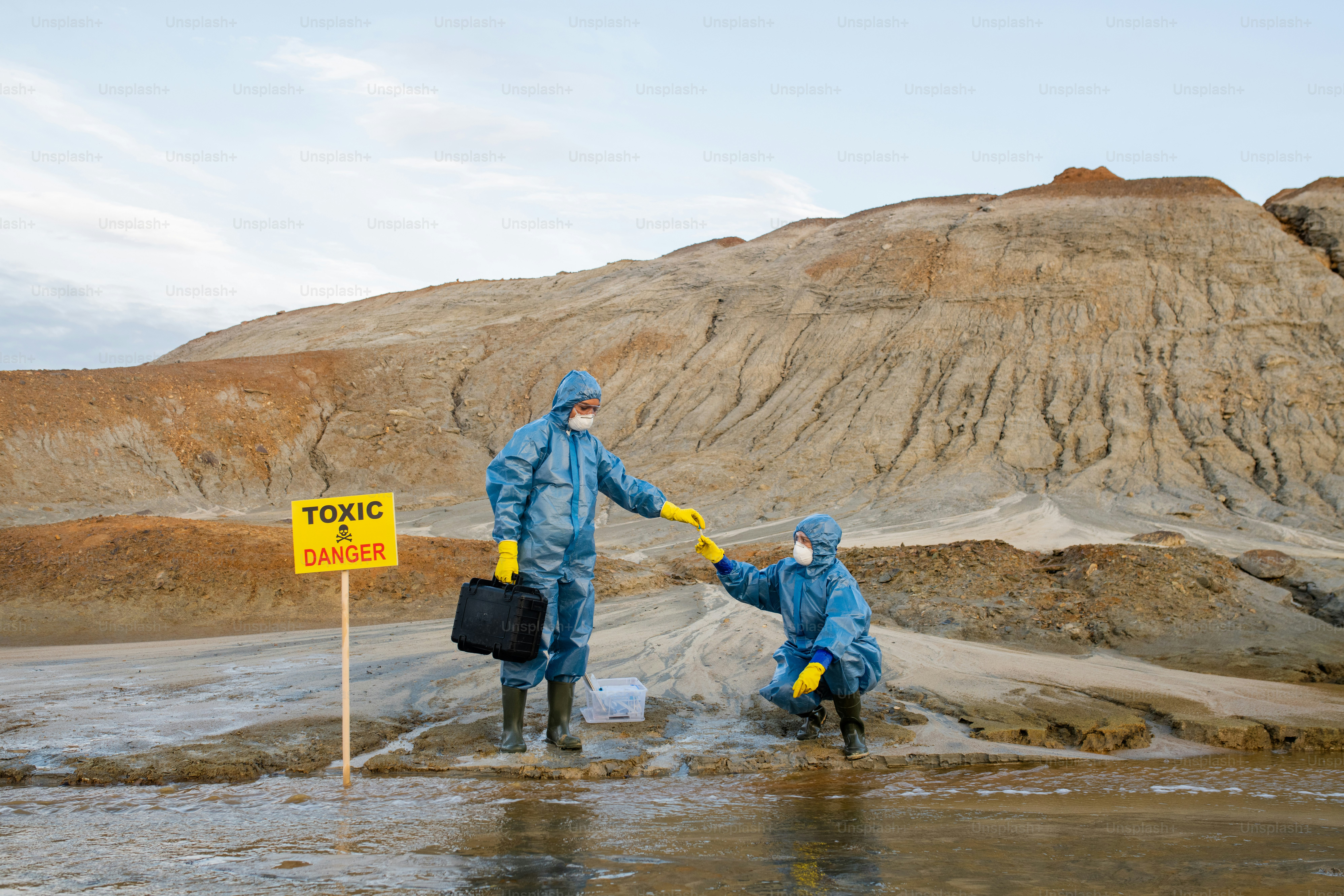 Ecologist in protective workwear taking sample of polluted water or soil from hand of colleague during scientific research in dangerous area