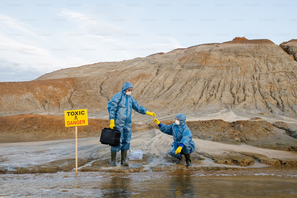 Ecologo in abbigliamento da lavoro protettivo che preleva un campione di acqua o terreno inquinato dalla mano del collega durante la ricerca scientifica in zona pericolosa