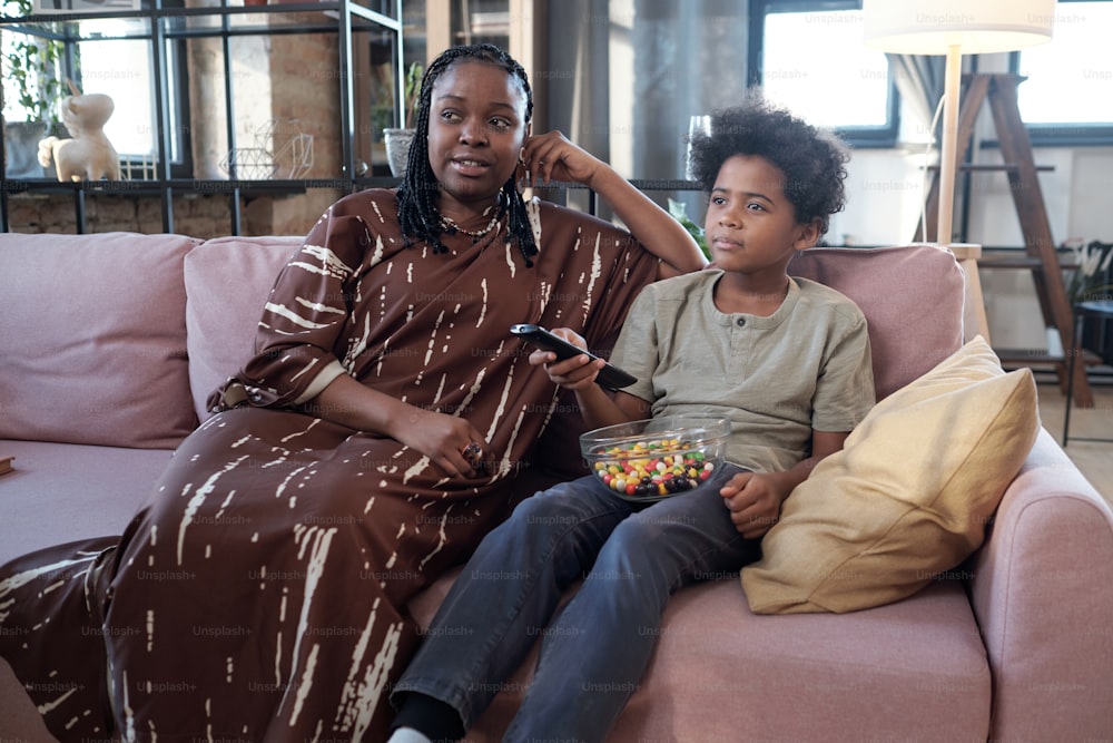 Young African female sitting next to her little son with bowl of candies while both resting on couch in living-room and watching tv