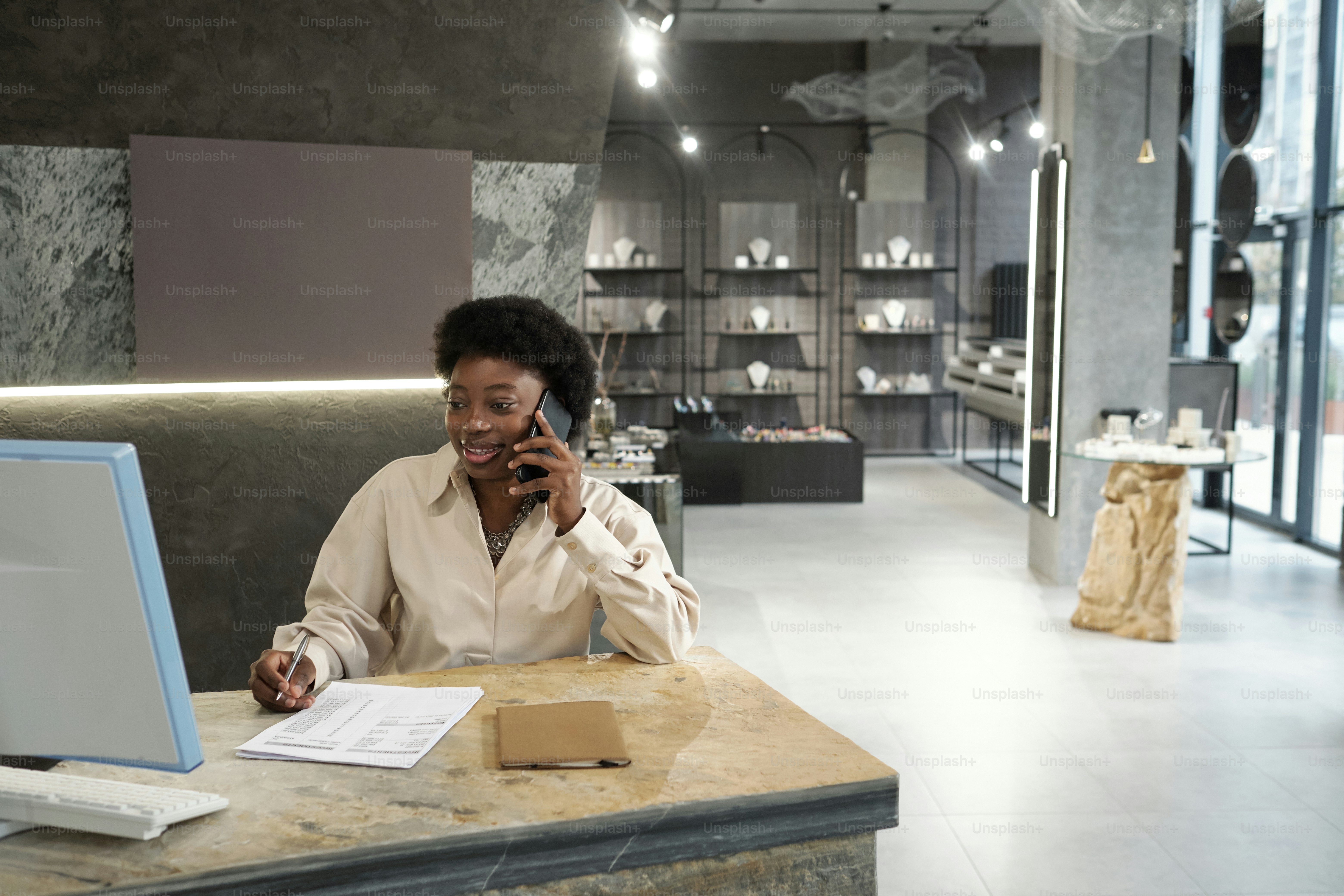 Young successful female owner of jewelry boutique consulting clients online and on mobile phone while sitting by workplace