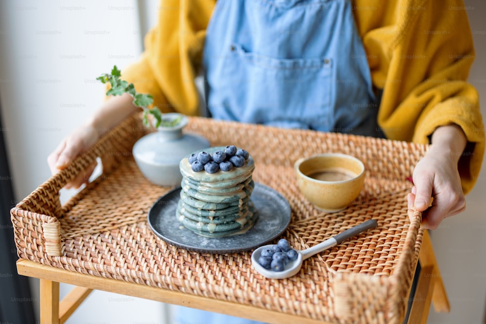 Woman in blue apron holding rattan tray with blue american pancakes with blueberry poured with caramel souse, cup of coffee and vase with flower. Homemade tasty food. Celebration of Shrovetide.