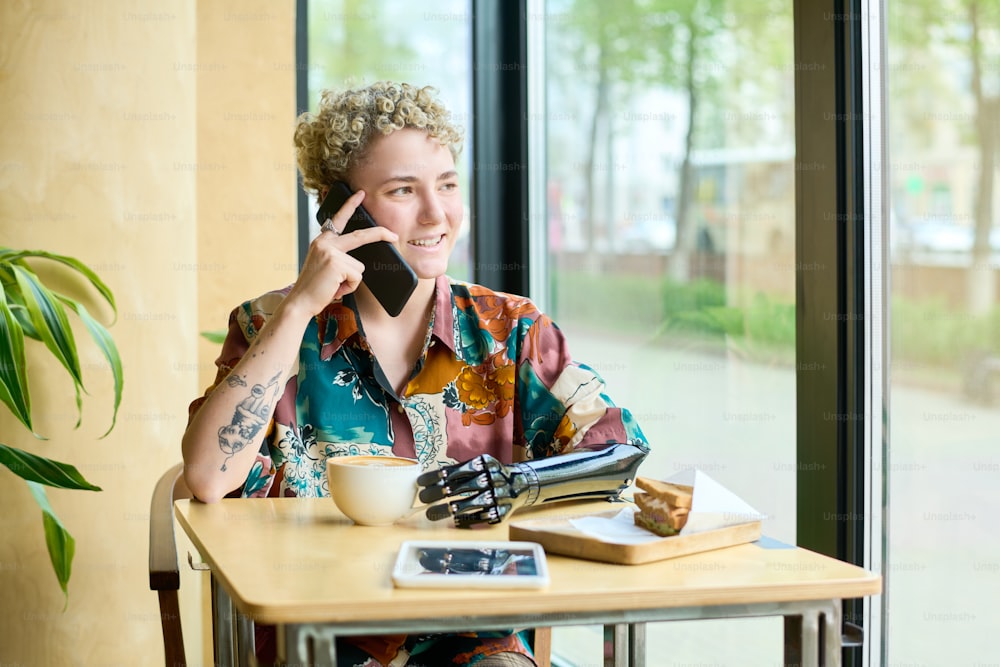Happy young female with partial arm speaking on mobile phone while sitting by table in cafe, looking through window and having coffee