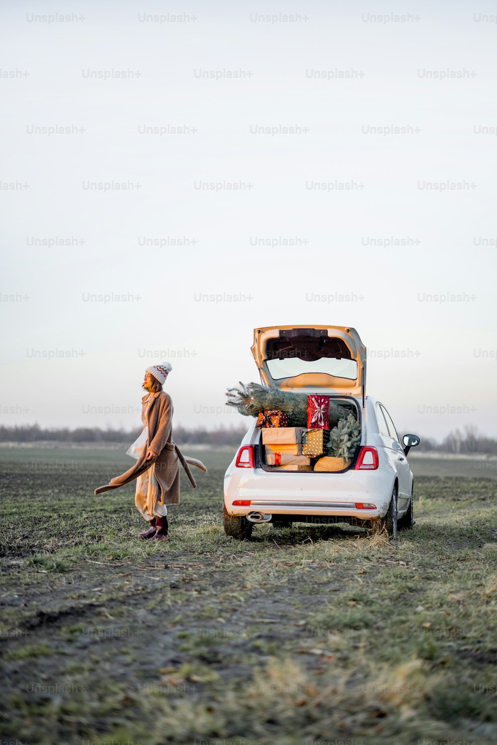 Woman in winter clothes walks near car full of giftboxes and Christmas tree on the field at sunset. Wide landscape with copy space on sky. Concept of New Year's mood
