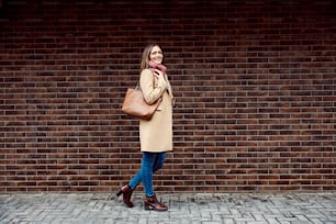 Beauty, fashion, and people concept. Full length of a happy fashionable woman walking on the street and passing by a brick wall.