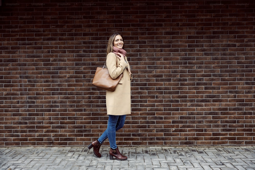 Beauty, fashion, and people concept. Full length of a happy fashionable woman walking on the street and passing by a brick wall.