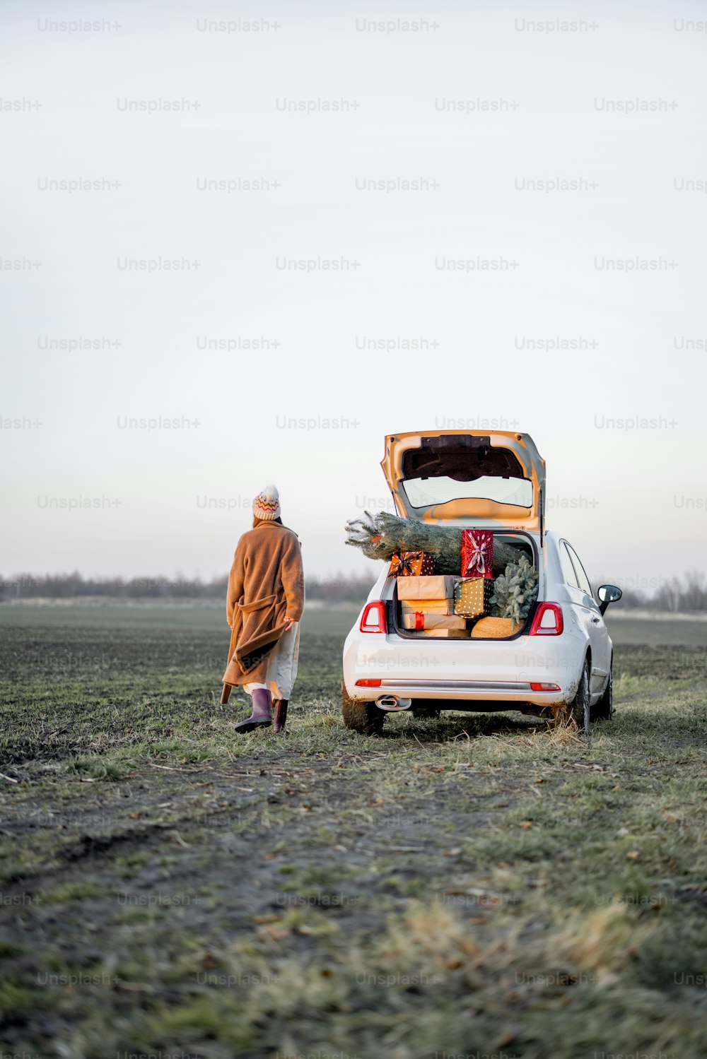 Woman in winter clothes walks near car full of giftboxes and Christmas tree on the field at sunset. Wide landscape with copy space on sky. Concept of New Year's mood