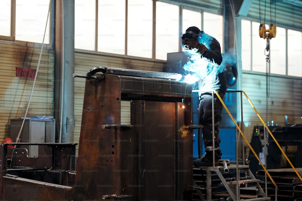 Contemporary worker or welder in protective mask and uniform using arc welding while repairing huge industrial machine in modern plant