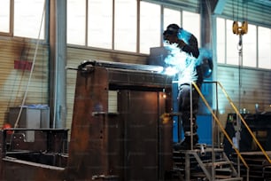 Trabajador o soldador contemporáneo con máscara protectora y uniforme que usa soldadura por arco mientras repara una enorme máquina industrial en una planta moderna