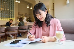 カフェのテーブルでコーヒーを飲むためにノートにメモを取る彼女の一日を計画するアジアの若いフリーランサー