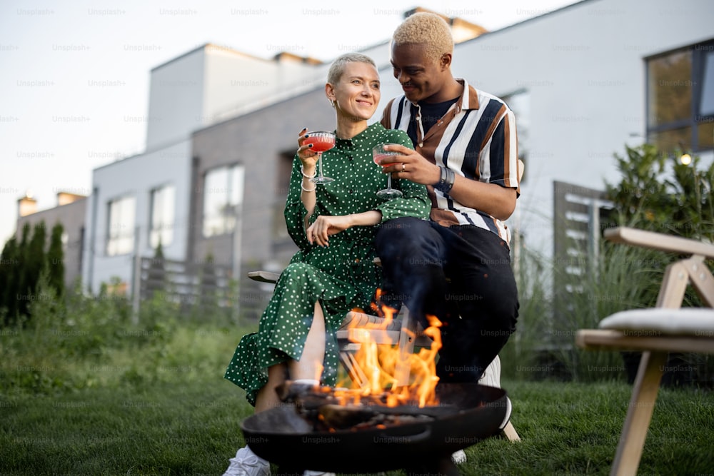 Multiracial couple drinking cocktails during home party in their garden. Burning fire for bbq. Concept of relationship. Idea of holiday or event. Black man and european girl enjoying time together