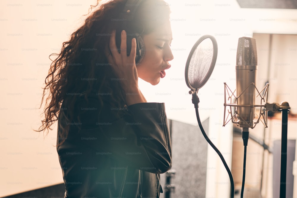 Ritratto di bella donna riccia in giacca di pelle e cappello che registra la voce in studio musicale su apparecchiature audio professionali, indossando cuffie