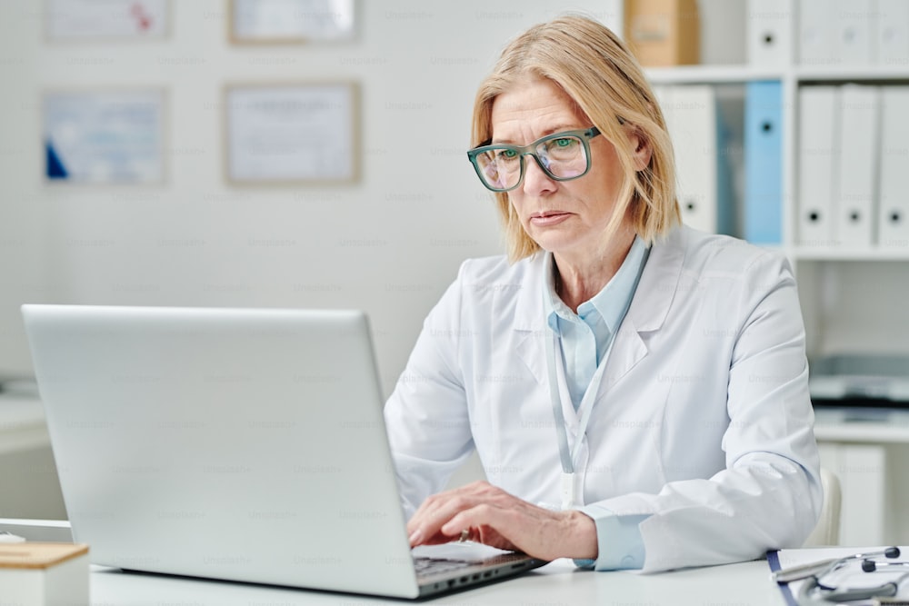Serious mature blond female doctor in whitecoat and eyeglasses typing on laptop keyboard while sitting by desk in medical office