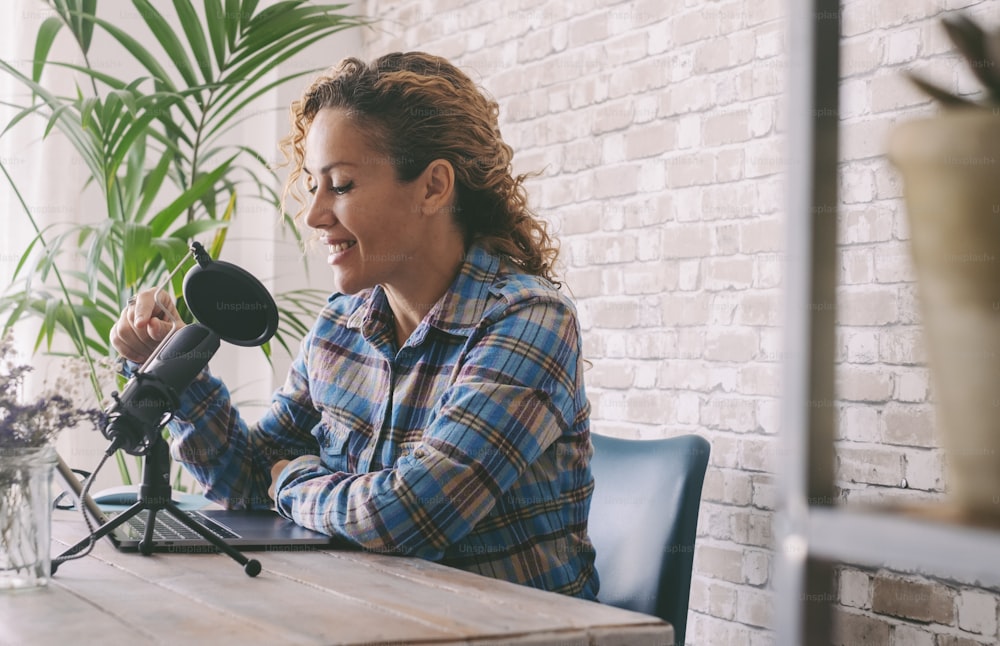 Young attractive woman recording podcast online on laptop sitting in home work place. Smart working and online modern job activity. Adult female at office with microphone. Alternative lifestyle work