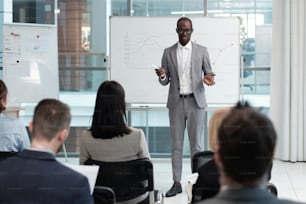 Young African American male coach explaining financial data to audience while standing by whiteboard with graphs in lecture hall