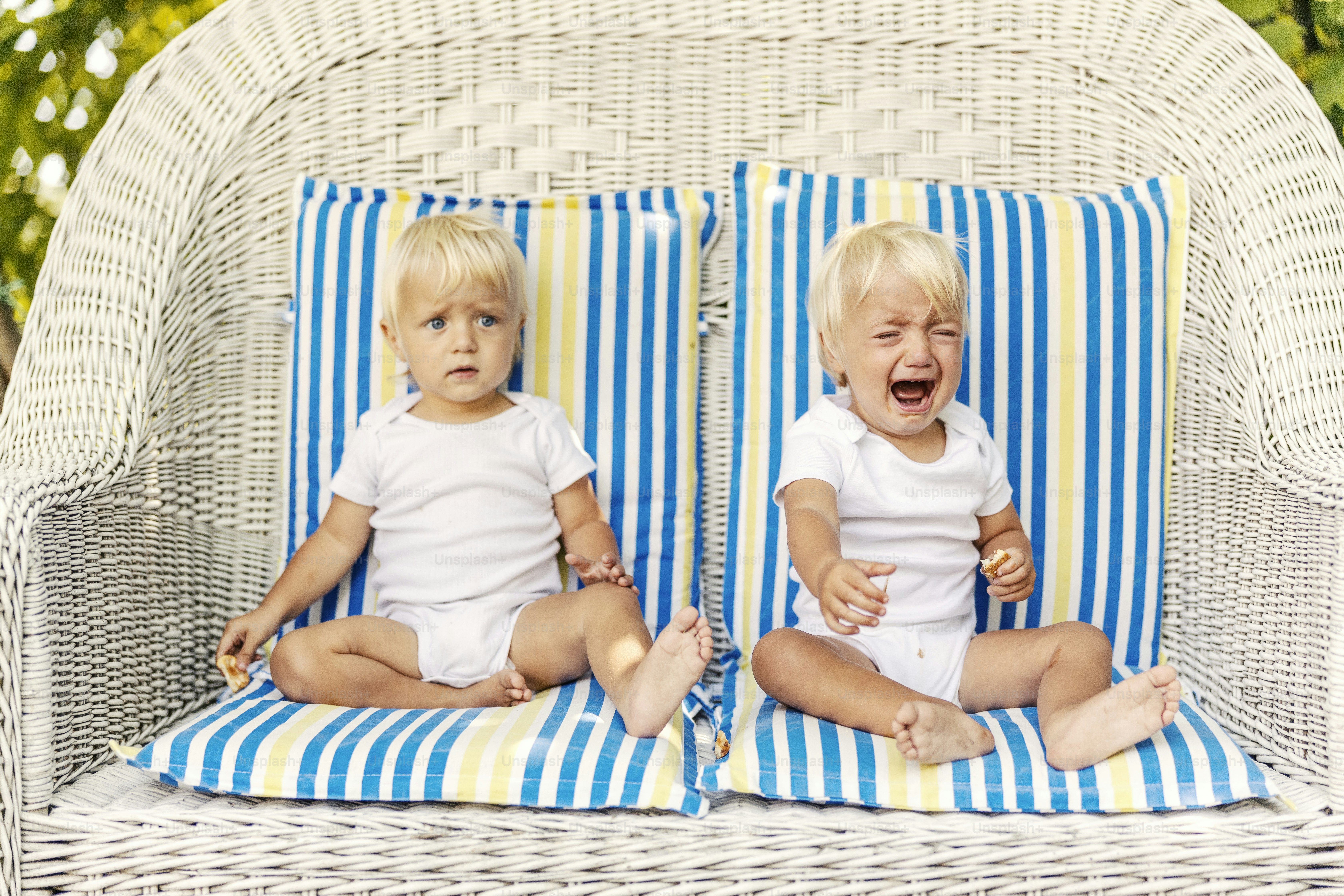 Toddler twins on a canvas chair with pillows Cute babies with blue eyes and blonde hair in white children's bodysuits sit on a huge outdoor wicker chair An anxious crying baby is looking for attention