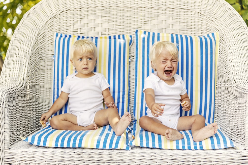 베개가 있는 캔버스 의자에 유아 쌍둥이 파란 눈과 흰색 어린이 바디 수트를 입은 금발 머리를 가진 귀여운 아기들이 거대한 야외 고리 버들 의자에 앉아 불안한 우는 아기가 관심을 찾고 있습니다.