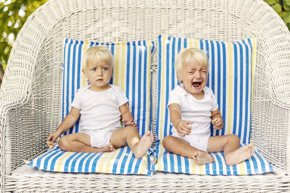 Gêmeos de criança em uma cadeira de lona com travesseiros Bebês bonitos com olhos azuis e cabelos loiros em bodysuits brancos de crianças sentam-se em uma enorme cadeira de vime ao ar livre Um bebê ansioso chorando está procurando atenção