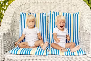 Gemelos pequeños en una silla de lona con almohadas Lindos bebés con ojos azules y cabello rubio en trajes blancos para niños se sientan en una enorme silla de mimbre al aire libre Un bebé ansioso que llora busca atención