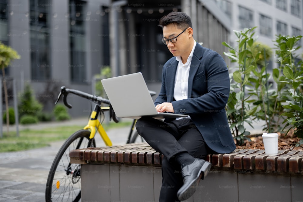 Hombre de negocios asiático que trabaja en una computadora portátil en la calle de la ciudad. Concepto de hombre exitoso moderno. Idea de trabajo remoto y freelance. Persona con gafas y traje. Hombre sentado en el banco