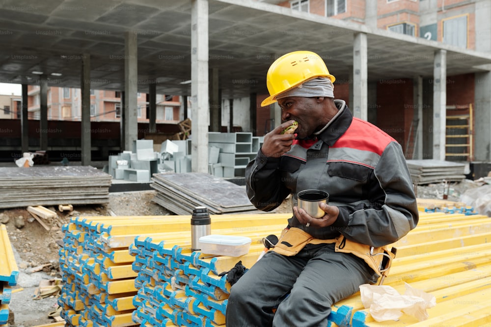 Hombre negro maduro hambriento con casco y ropa de trabajo comiendo sándwich y bebiendo té caliente mientras está sentado en una pila de materiales de construcción