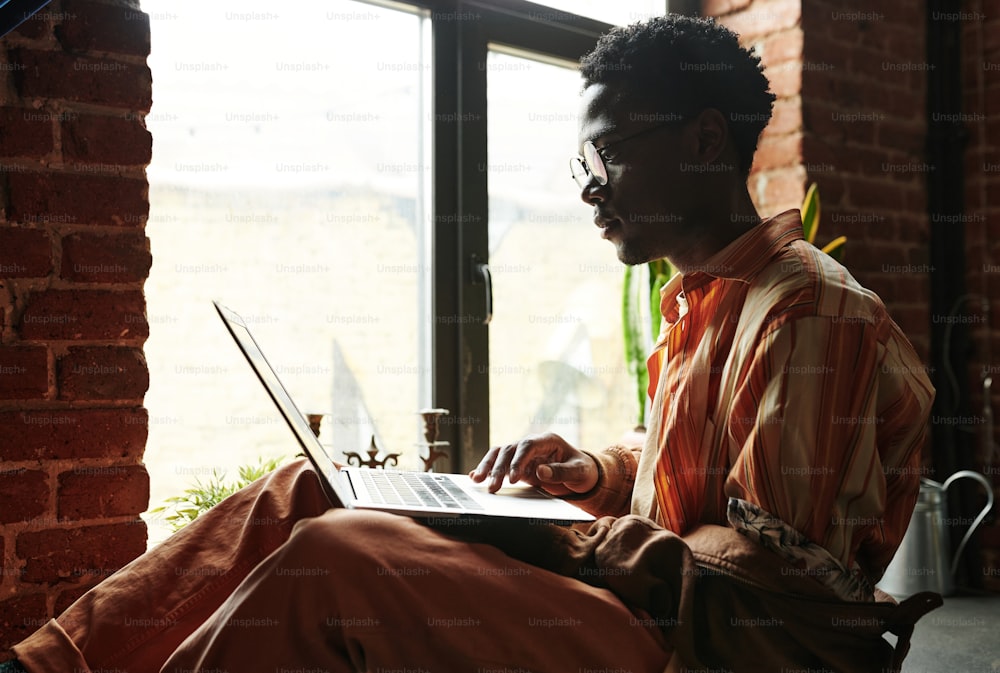 Side view of young black man looking at laptop screen while sitting against window in loft apartment and networking