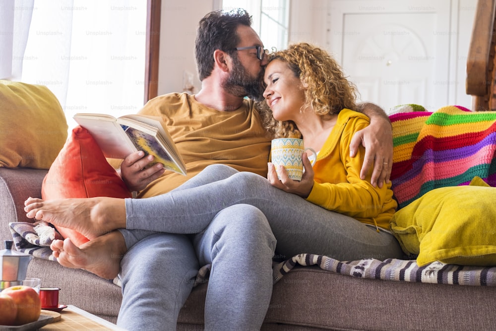 Happy beautiful caucasian couple at home enjoy morning leisure activity in relax together - love and relationship life with young people sitting on the colourful sofa with book and coffee