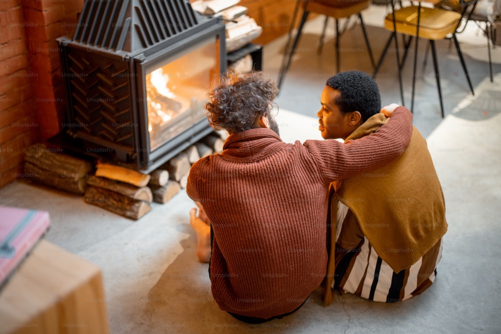 Two men sitting together by the burning fireplace at cozy home. Concept of homosexual relations and coziness on winter time. Idea of multinational gay families. View from the backside
