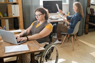 Young disable businesswoman sitting in wheelchair in front of laptop and scrolling through online data on background of her busy colleague